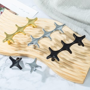 May include: A collection of star-shaped cabinet pulls in gold, silver, and black. The hardware is displayed on a wooden board. The pulls have a modern design, with four points extending from a central hub. The brand name "JANE HARDWARE" is visible.