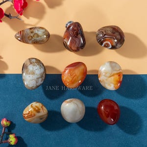 Könnte beinhalten: Eine Sammlung polierter Achatsteine in verschiedenen Formen und Farben, darunter Braun, Orange und Weiß. Die Steine werden auf einem zweifarbigen Hintergrund mit dem Text "JANE HARDWARE" präsentiert.