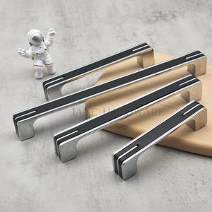 Op de afbeelding: Vier zilveren kastgrepen met zwarte accenten worden getoond op een houten plank. De grepen hebben een rechthoekige vorm met een centrale zwarte streep en zilveren strepen. Een kleine astronautfiguur staat op de achtergrond.