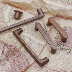 Könnte beinhalten: Eine Sammlung von Holzschrankbeschlägen, darunter Griffe und ein Knauf, in verschiedenen Größen. Die Beschläge sind aus dunkelbraunem Holz gefertigt und haben eine glatte, polierte Oberfläche. Die Griffe sind rechteckig mit gebogenen Enden.