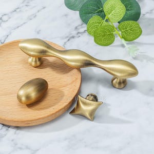 Może przedstawiać: Zestaw trzech ceramicznych kubków z nakrapianą, kremową glazurą. Każdy kubek ma inny ręcznie malowany wzór: niebiesko-zielony liść, żółte słońce i różowy kwiat. Kubki mają prosty, nowoczesny design.