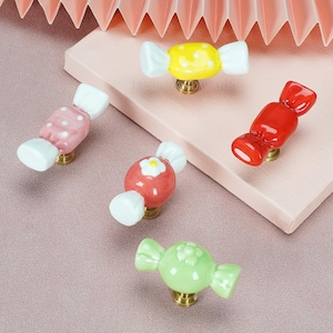 May include: Five candy-shaped cabinet knobs with brass-colored bases. The set includes a yellow knob with white dots, a pink knob with white dots, a solid red knob, a red knob with a white flower, and a solid light green knob with a flower detail.