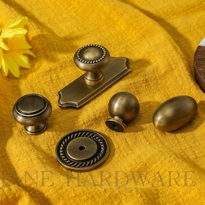 Puede incluir: Un surtido de herrajes de gabinete de latón antiguo, que incluye perillas, un tirador y una placa trasera, dispuestos sobre tela amarilla. El herraje tiene una estética vintage, con diseños detallados y un acabado cálido y envejecido. El texto "JANE HARDWARE" es visible.