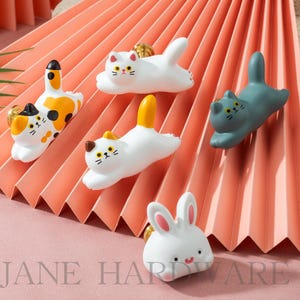 May include: A collection of decorative cabinet knobs in the shape of cats and a bunny. The knobs are white, orange, and gray with gold-colored hardware. The text "JANE HARDWARE" is visible at the bottom of the image.