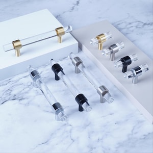 Może przedstawiać: Przezroczyste akrylowe uchwyty do szafek ze złotym, srebrnym i czarnym osprzętem. Uchwyty mają cylindryczną konstrukcję i są ułożone na białej i szarej powierzchni. Okucia są w różnych metalicznych wykończeniach.