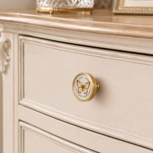 May include: A round cabinet knob with a brushed gold metal finish and a circular white center insert. The insert features a detailed illustration of a yellow and orange butterfly surrounded by a delicate green leaf wreath design.