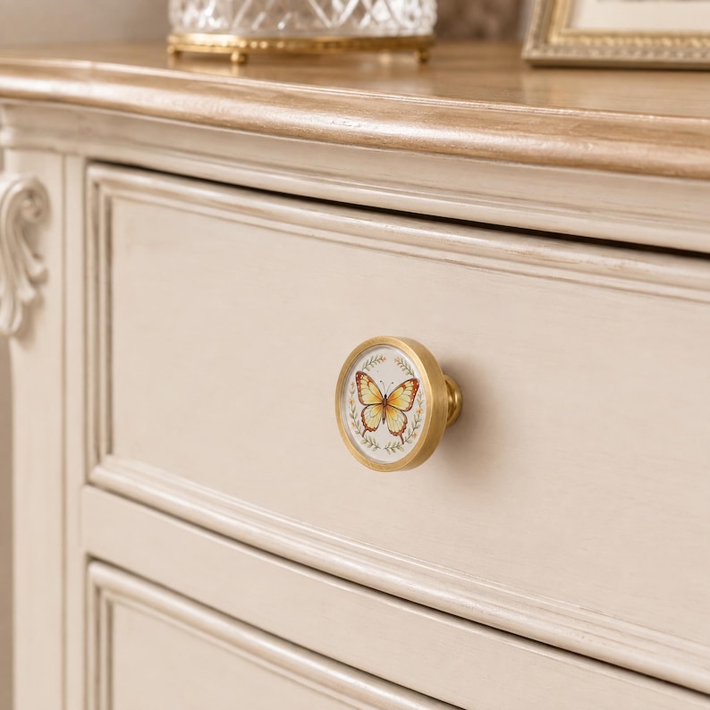 May include: A round cabinet knob with a brushed gold metal finish and a circular white center insert. The insert features a detailed illustration of a yellow and orange butterfly surrounded by a delicate green leaf wreath design.