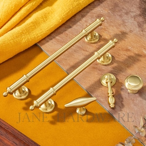 May include: A collection of gold-toned cabinet hardware, including two long bar pulls, a small knob, a handle, and a decorative pull. The hardware is arranged on a yellow and brown surface. The text "JANE HARDWARE" is visible.