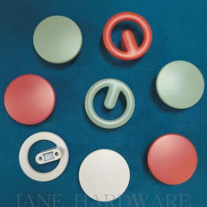 Puede incluir: Una colección de piezas de ferretería coloridas, que incluyen componentes redondos y en forma de anillo en rojo, verde y blanco. El texto "JANE HARDWARE" está impreso en la parte inferior. Las piezas están dispuestas sobre una superficie azul.