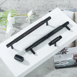 May include: A collection of black cabinet hardware, including handles and a knob, with a textured, grooved design. The hardware is displayed on a white surface, with the text "JANE HARDWARE" visible. The hardware is designed for cabinets and drawers.