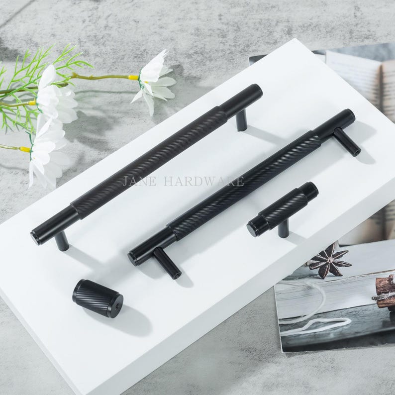May include: A collection of black cabinet hardware, including handles and a knob, with a textured, grooved design. The hardware is displayed on a white surface, with the text "JANE HARDWARE" visible. The hardware is designed for cabinets and drawers.