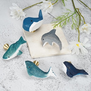 May include: Five drawer knobs shaped like marine animals, including a dark blue whale, a grey dolphin, a teal shark, a teal whale, and a dark blue whale. Each knob features a gold-toned metal base and a faceted, carved wooden surface.