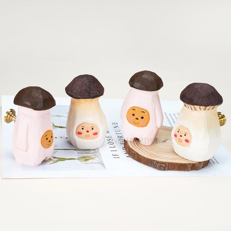 Op de afbeelding: Vier handgesneden houten ladeknoppen in de vorm van paddenstoelen met donkerbruine hoeden en lichtroze of cr&egrave;mekleurige stelen. Elke steel heeft een geschilderd cartoongezicht met roze wangen. Twee knoppen hebben goudkleurig metalen beslag aan de zijkanten.