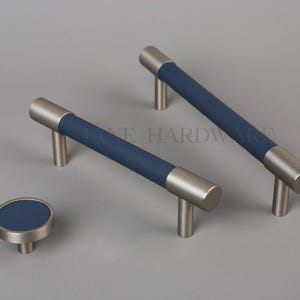 Puede incluir: Un juego de herrajes para armarios con dos tiradores de barra y un pomo redondo. Los tiradores tienen un acabado de metal cepillado con un centro azul marino. El pomo tiene un centro azul marino a juego. Los herrajes se muestran sobre una superficie gris.