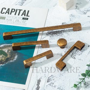 May include: A collection of antique brass hardware, including cabinet pulls and a knob, arranged on a magazine. The hardware has a weathered finish and a rectangular design. The magazine's title is "CAPITAL" with the word "HARDWARE" visible.