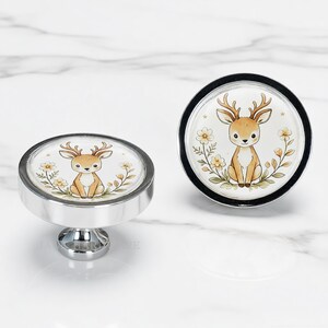May include: Two round cabinet knobs with a polished silver metal finish. The center of each knob features a circular illustration of a light brown deer with antlers, surrounded by small yellow flowers and green leaves on a white background.