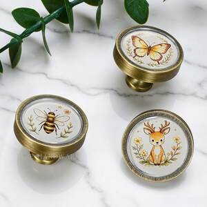 May include: Three round cabinet knobs with an antique brass finish. Each knob features a circular insert with a different illustration: a yellow butterfly, a black and yellow bee, and a brown deer, all surrounded by delicate floral leaf motifs.