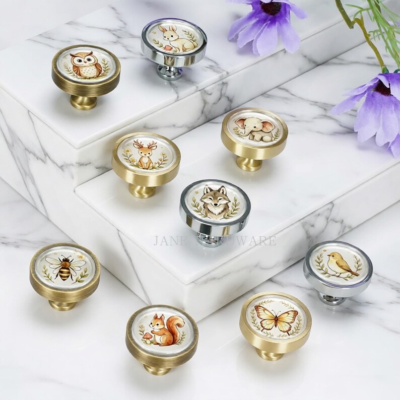 May include: A collection of nine round cabinet knobs featuring various animal illustrations including an owl, rabbit, deer, elephant, fox, bee, squirrel, bird, and butterfly. Each knob has a metallic base and a circular top with a printed design.