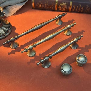 Può includere: Set di ferramenta per mobili in bronzo antico. Il set comprende tre maniglie lunghe e due pomelli. Le maniglie hanno un design curvo con estremità decorative. I pomelli sono rotondi con un bordo dettagliato. L'hardware è esposto su una superficie marrone-rossastra.