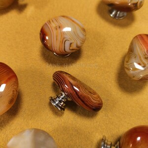 May include: A collection of decorative cabinet knobs made from polished stone. The knobs feature swirling patterns in shades of brown, orange, and white. Each knob is mounted on a silver metal base. The knobs are displayed on a yellow-tan surface.