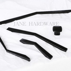 Może przedstawiać: Zestaw czarnych okuć do szafek, w tym uchwyty i gałka, na białym tle. Uchwyty mają nowoczesny, kątowy design. Widoczny jest tekst "JANE HARDWARE".