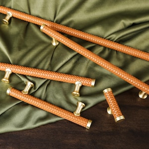 May include: A collection of brown leather cabinet pulls with a woven texture and gold-toned hardware. The pulls are arranged on a dark wooden surface with a green satin fabric backdrop. The hardware is a gold color.