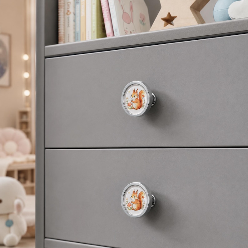 May include: Two circular drawer knobs with a silver-toned metal rim and a central white inlay featuring a colorful illustration of a squirrel standing next to a small mushroom. The knobs are mounted on a smooth, matte gray wooden drawer front.