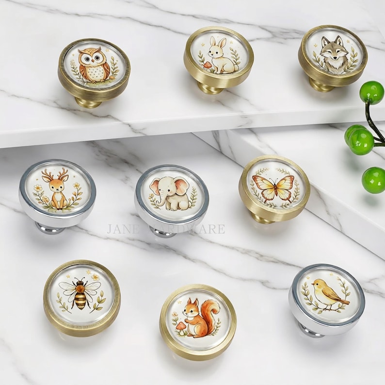 May include: A collection of nine round cabinet knobs featuring watercolor illustrations of animals including an owl, rabbit, fox, deer, elephant, butterfly, bee, squirrel, and bird. The knobs have metallic gold or silver finishes and circular designs.