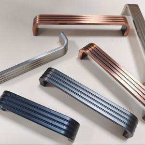 May include: An assortment of cabinet pulls in various metallic finishes, including copper, silver, and black. Each pull features a curved design with horizontal grooves, offering a modern aesthetic. The pulls are displayed on a light surface.