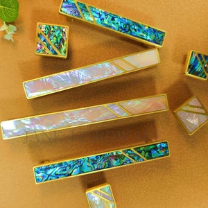 Könnte beinhalten: Eine Sammlung dekorativer Schrankbeschläge, darunter rechteckige Griffe und quadratische Knöpfe. Die Beschläge verfügen über schillernde Abalone-Muschel-Inlays mit goldfarbenen Metallrahmen. Die Griffe variieren in der Länge, und die Knöpfe sind quadratisch.