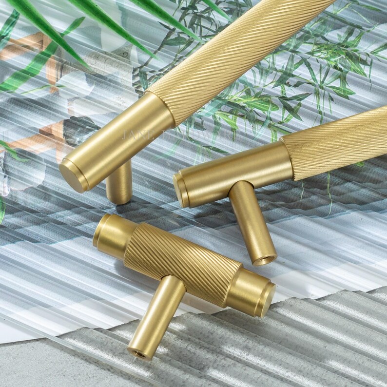 May include: Close-up of three gold-colored cabinet pulls. The pulls have a cylindrical design with a textured, ridged pattern on the handle. The T-shaped base is smooth and solid. The background is a mix of green and gray.