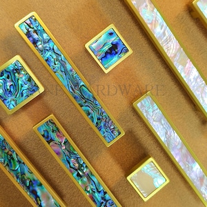 Könnte beinhalten: Eine Sammlung goldfarbener Schrankbeschläge, darunter rechteckige Griffe und quadratische Knöpfe. Die Beschläge verfügen über schillernde Abalone-Muschel-Inlays mit blauen, grünen und violetten Farbtönen. Die Beschläge sind auf einem neutralen Hintergrund angeordnet.