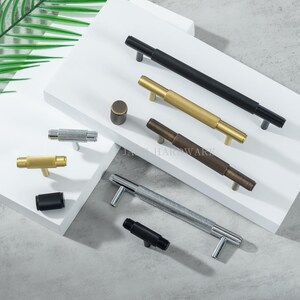 May include: A collection of cabinet hardware, including black, gold, silver, and bronze-toned handles and knobs. The handles have a textured, ridged design. The knobs are cylindrical. The hardware is displayed on a white surface.