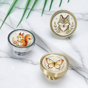 May include: Three round cabinet knobs featuring illustrations of a squirrel with a mushroom, a fox surrounded by leaves, and a butterfly. The knobs have metallic finishes in silver, antique brass, and brushed gold, each displaying a nature-themed design.