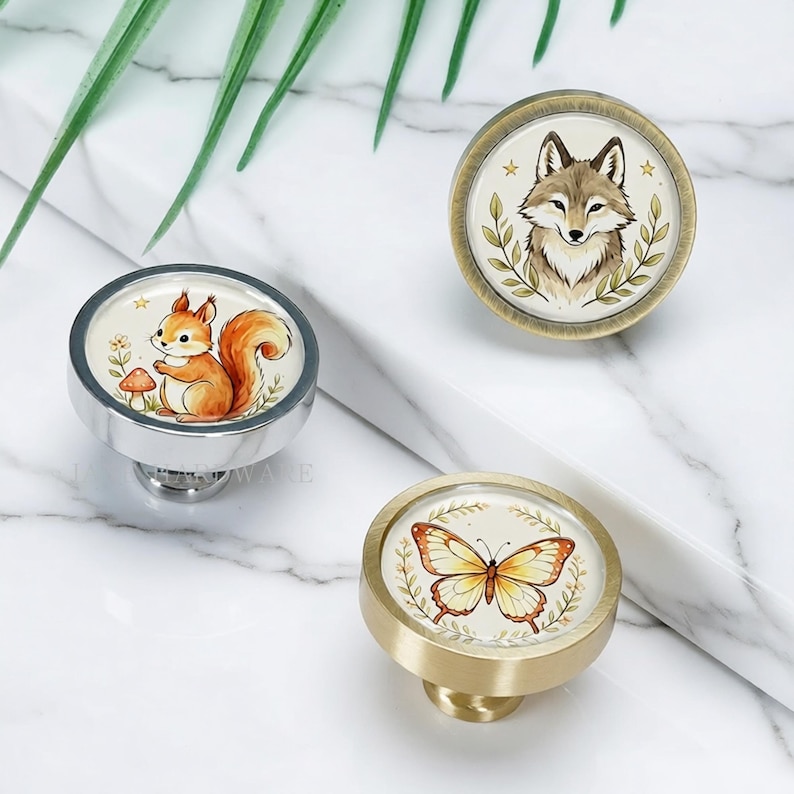 May include: Three round cabinet knobs featuring illustrations of a squirrel with a mushroom, a fox surrounded by leaves, and a butterfly. The knobs have metallic finishes in silver, antique brass, and brushed gold, each displaying a nature-themed design.