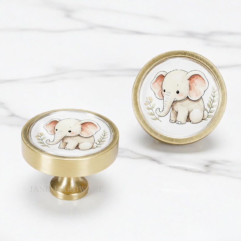 May include: Two round cabinet knobs with a brushed brass finish. Each knob features a circular insert displaying a watercolor-style illustration of a small, light-colored elephant with pink ears, surrounded by delicate green leafy sprigs.