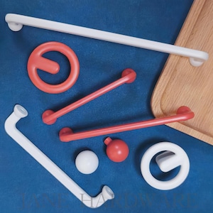 Könnte beinhalten: Eine Auswahl an roten und weißen Badezimmer-Sicherheitszubehör, darunter Haltegriffe, ein kreisförmiger Griff und ein gebogener Griff. Die Artikel sind auf einer blauen Oberfläche angeordnet, mit einem Holztablett im Hintergrund.