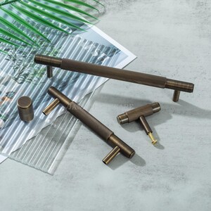 May include: A collection of bronze-toned cabinet hardware, including a long bar pull, two T-shaped pulls, and a cylindrical knob. The hardware features a textured, ridged design. The items are arranged on a light gray surface with a leaf and glass accents.