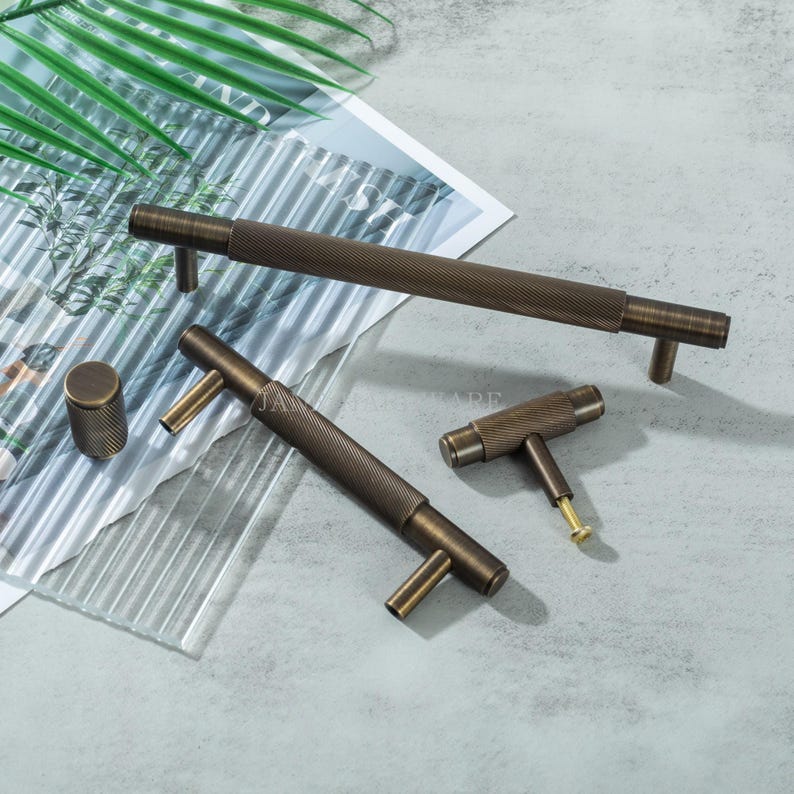 May include: A collection of bronze-toned cabinet hardware, including a long bar pull, two T-shaped pulls, and a cylindrical knob. The hardware features a textured, ridged design. The items are arranged on a light gray surface with a leaf and glass accents.