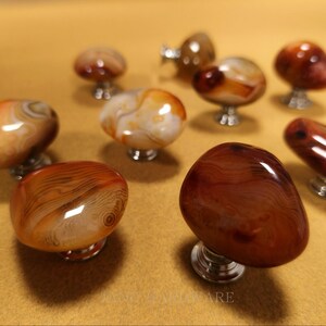 May include: A selection of polished agate cabinet knobs in brown, orange, and cream hues. Each knob features a distinct pattern and is mounted on a silver-coloured base. The knobs are displayed on a light brown surface.