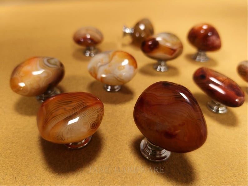 May include: A selection of polished agate cabinet knobs in brown, orange, and cream hues. Each knob features a distinct pattern and is mounted on a silver-coloured base. The knobs are displayed on a light brown surface.
