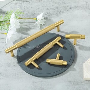 May include: A collection of gold-colored cabinet hardware, including long bar pulls and T-shaped knobs, with a textured, ridged design. The hardware is arranged on a gray surface, with the text "JANE HARDWARE" visible. White flowers and greenery are in the background.