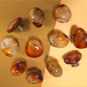 May include: A selection of agate stone cabinet knobs in different shapes and sizes. The knobs display warm colours, including orange, brown, and cream, with unique patterns. Each knob has a silver-coloured base for mounting.