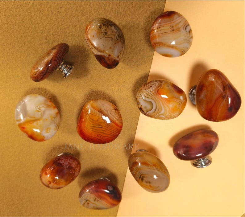 May include: A selection of agate stone cabinet knobs in different shapes and sizes. The knobs display warm colours, including orange, brown, and cream, with unique patterns. Each knob has a silver-coloured base for mounting.