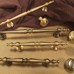 Puede incluir: Una variedad de herrajes de gabinete de latón antiguo, que incluyen manijas y perillas de varios tamaños. Las manijas tienen detalles ornamentados y las perillas son redondas con patrones decorativos.