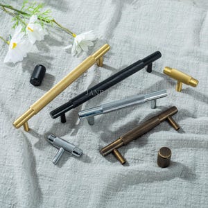 May include: A collection of decorative cabinet hardware in various metallic finishes, including gold, black, silver, and bronze. The hardware features textured, ridged designs and includes both pulls and knobs. The arrangement is on a neutral-toned fabric surface.