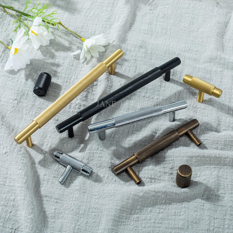 May include: A collection of decorative cabinet hardware in various metallic finishes, including gold, black, silver, and bronze. The hardware features textured, ridged designs and includes both pulls and knobs. The arrangement is on a neutral-toned fabric surface.