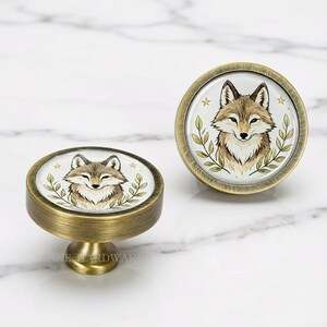 May include: Two round cabinet knobs with an antique brass finish. The center of each knob features a circular illustration of a brown and white fox head surrounded by green leafy branches and small yellow stars on a white background.