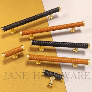 以下が含まれることがあります： 黒と茶色の編み革のハンドルが付いたキャビネットプル。プルは金色のベースとエンドキャップが付いています。画像はニュートラルな背景に設定されており、ハードウェアのデザインと質感を際立たせています。