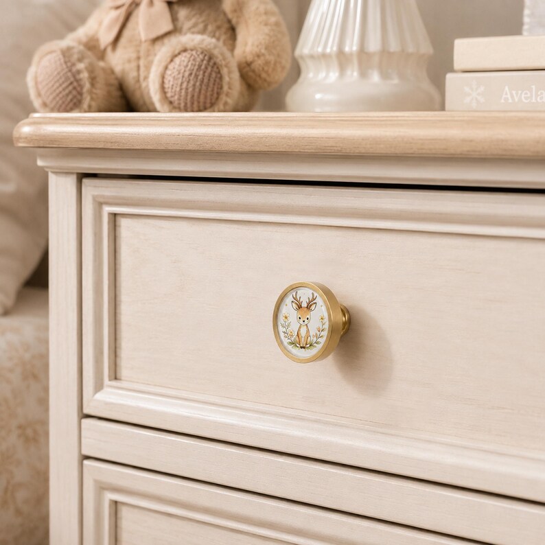 May include: A round drawer knob with a brushed gold metal finish featuring a circular insert. The insert displays a detailed illustration of a small brown deer with antlers, surrounded by a wreath of green leaves and small yellow flowers.